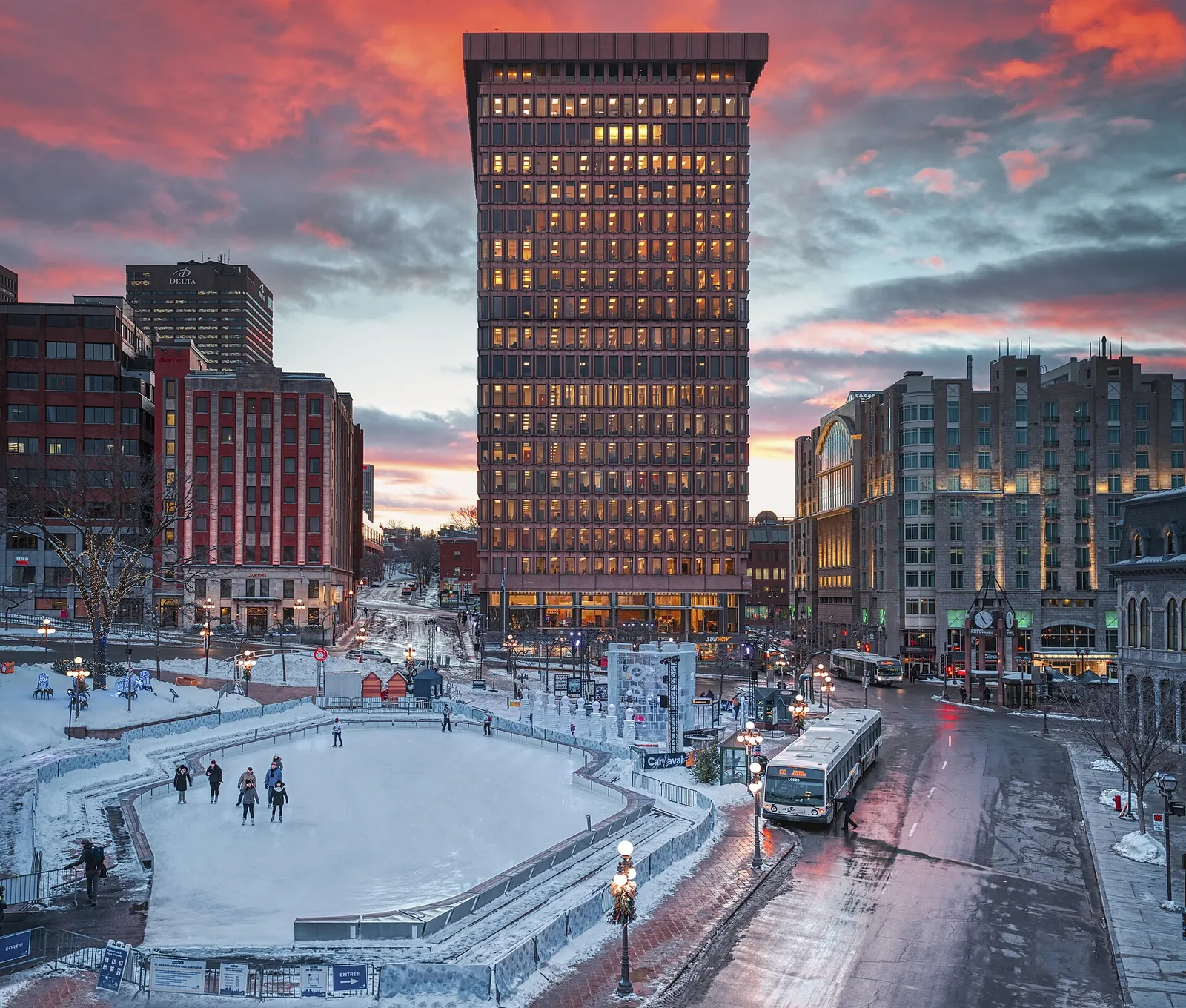 Skaters on the outdoor rink at Place D'Youville in Quebec City at dusk, with a tall art deco tower behind